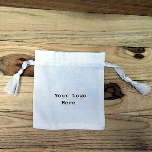 May include: White cotton drawstring bag with a white tassel on each side. The bag has the text "Your Logo Here" printed on the front.