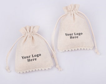May include: Two white cotton drawstring bags with pom pom trim. Each bag has the text "Your Logo Here" printed on the front.
