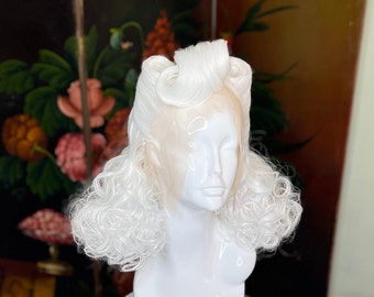 May include: A white wig with a vintage hairstyle is displayed on a white mannequin head. The wig features voluminous, curly hair and a styled top section. The mannequin is set against a backdrop of a floral painting.