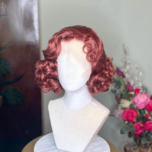 May include: A vintage-style auburn wig with short, curly hair. The wig is styled with finger waves and curls, and is displayed on a white mannequin head. The wig is a reddish-brown color and appears to be made of synthetic fibers.