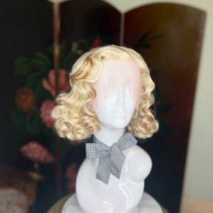 May include: A blonde wig with a light brown highlight, styled in a vintage, curled bob. The wig is on a white mannequin head with a black and white gingham bow.