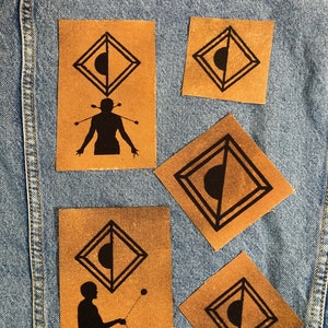 Op de afbeelding: Vier bruine stoffen patches met zwarte ontwerpen. De patches hebben een geometrische vorm met een halve cirkel erin. Twee van de patches hebben een silhouet van een persoon die jongleert.