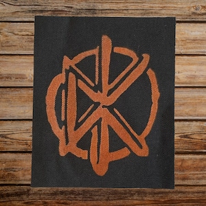 May include: A black fabric patch with an orange stylized logo. The logo is a circle with a cross inside. The cross is made up of four lines that intersect in the center of the circle.