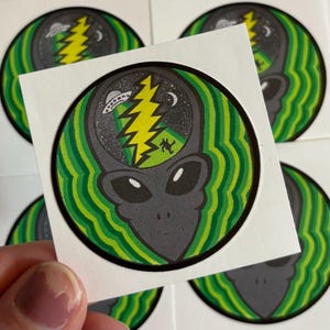 May include: A sticker featuring a gray alien head with a green and yellow design. The alien's head contains a starry sky, a lightning bolt, a UFO, and a dancing figure. The sticker has a green and white wavy border.