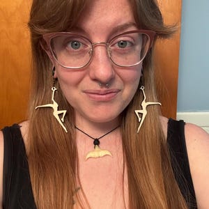 Op de afbeelding: Een vrouw draagt een bril met gouden montuur, een zwarte tanktop en een ketting met een haaientandhanger. Ze draagt ook oorbellen met een danseresontwerp. Haar haar is lang en rossig.