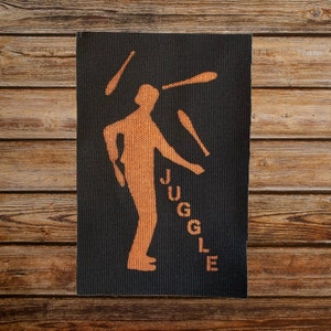 Op de afbeelding: Een zwarte patch met een oranje silhouet van een persoon die met drie keulen jongleert. Het woord "JUGGLE" staat verticaal in oranje letters aan de rechterkant van de patch.