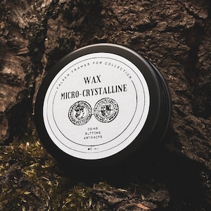 Könnte beinhalten: Eine schwarze runde Dose mit einem weißen Etikett, auf dem "Wax Micro-Crystalline" und "Coins Buttons Artifacts" steht. Die Dose steht auf einem mit Moos bewachsenen Baumstamm.