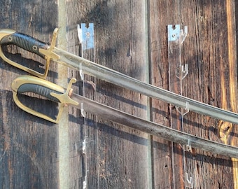 Puede incluir: Dos espadas montadas en una pared de madera con soportes de espada de plástico transparente. Las espadas tienen detalles dorados y plateados. El texto "CK36" es visible en la esquina inferior izquierda de la imagen.