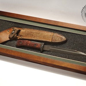 May include: A framed display of a vintage military knife and its leather sheath. The knife has a wooden handle and a metal blade. The sheath is brown leather with stitching and a metal snap.