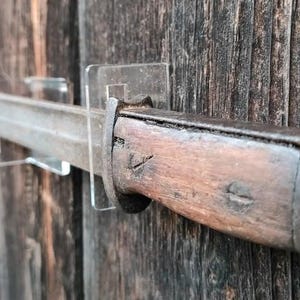 Op de afbeelding: Een vintage bajonet met een houten handvat en een metalen blad is gemonteerd op een houten muur met transparante acryl displaybeugels. Het bajonet heeft een metalen ring in de buurt van het handvat en de tekst "CK38" is zichtbaar op de muur.
