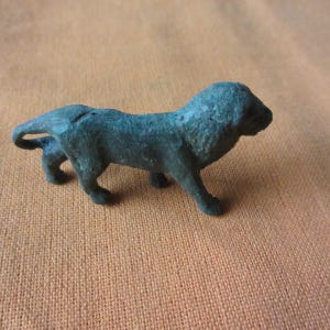 Puede incluir: Pequeña figura de león verde oscuro. El león está en posición de caminar, con la cabeza ligeramente girada hacia la derecha. La figura tiene una superficie texturizada y está sobre un fondo marrón.