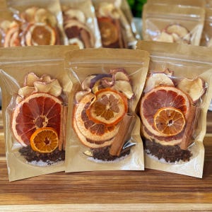May include: Clear plastic bags filled with dried orange and apple slices, cinnamon sticks, and cloves. The bags are sealed and arranged on a wooden surface. The dried fruit slices are orange and red in color.