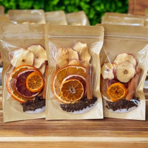 May include: Three clear plastic bags filled with dried fruit and spices. Each bag contains apple slices, orange slices, a cinnamon stick, and cloves. The bags are sealed and arranged on a wooden surface, with a green background.