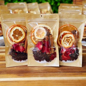 May include: Three clear, sealed bags filled with dried orange slices, cinnamon sticks, and red rose petals. The bags are arranged on a wooden surface, suggesting a natural product or a craft item.