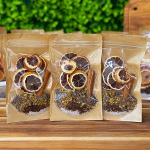 May include: Three clear, sealed pouches filled with dried orange slices, cinnamon sticks, and various herbs. The pouches are arranged on a wooden surface, with a green, blurred background. The contents appear to be a blend for a beverage.