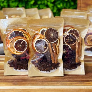 May include: Three clear plastic bags filled with dried orange slices, cinnamon sticks, and other botanicals. The bags are sealed and arranged in a row, with a wooden surface in the foreground and a blurred green background.
