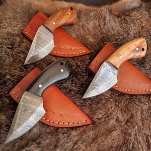 Puede incluir: Tres cuchillos hechos a mano con hojas de acero con patrones y mangos de madera. Cada cuchillo está en una funda de cuero marrón con costuras decorativas.