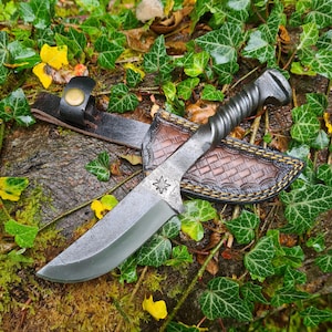 May include: A hand-forged steel knife with a twisted handle and a leather sheath. The blade has a starburst design. The sheath is brown with a woven pattern and a black leather strap with a brass button. The knife rests on a mossy log surrounded by green ivy.