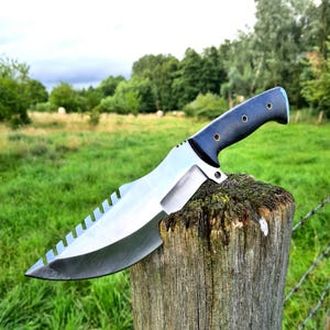 Könnte beinhalten: Ein großes Messer mit silberner Klinge, gezahntem Rand und dunkelblauem Griff. Das Messer liegt auf einem verwitterten Holzpfosten, mit einer grünen Wiese und Bäumen im Hintergrund. Die Klinge hat eine scharfe Spitze und ein gebogenes Design.