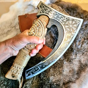 Könnte beinhalten: Eine Handaxt mit einer dunklen Metallklinge, verziert mit silberfarbenen Schnörkeln und einem Vegvisir-Symbol. Der Griff besteht aus hellem Holz mit geschnitzten geometrischen Mustern, Messingnieten und einer Umwicklung aus Jutekordel.