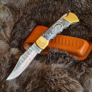 Peut inclure: Un couteau pliant avec une lame à motifs et un manche avec un grain de bois gris et des accents en laiton. Le couteau est présenté avec un étui en cuir marron, sur fond de fourrure brune.