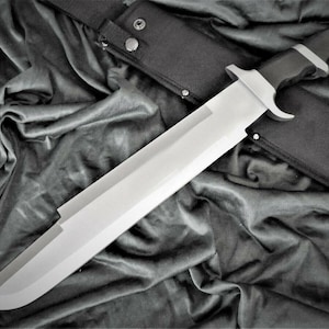 May include: A large, silver-colored knife with a black handle and a black sheath. The knife is lying on a black fabric background.