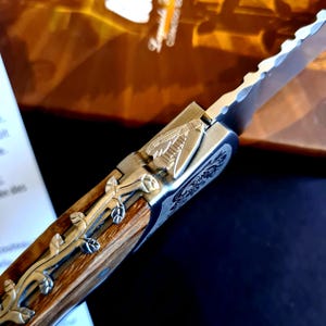 May include: Close-up of a partially open pocket knife. The knife features a wooden handle with silver floral embellishments and a serrated blade. The metal components have intricate engraved designs. The knife is set against a blurred background.