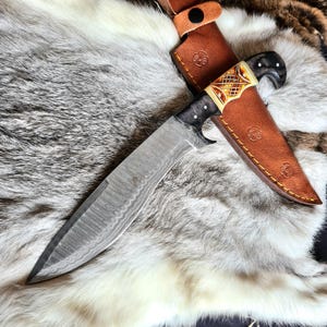 May include: A large, ornate knife with a patterned blade and a decorative handle. The handle features a combination of black, cream, and gold-colored elements. It rests on a fur-like surface, with a brown leather sheath.