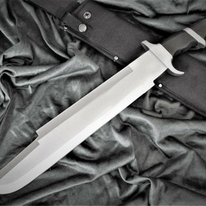 May include: A large, silver-colored knife with a black handle and a black sheath. The knife is lying on a black fabric background.