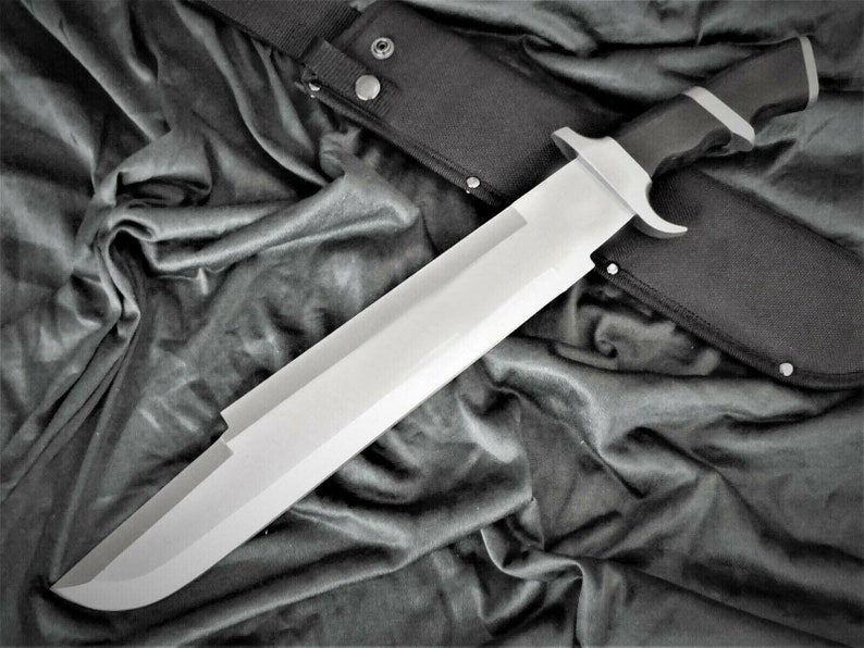 May include: A large, silver-colored knife with a black handle and a black sheath. The knife is lying on a black fabric background.