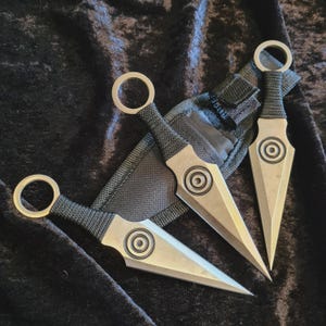Può includere: Tre coltelli da lancio argentati con manici avvolti in nero e anelli circolari per le dita. Ogni coltello presenta un design circolare centrale. I coltelli sono esposti con un fodero nero su una superficie di velluto scuro.