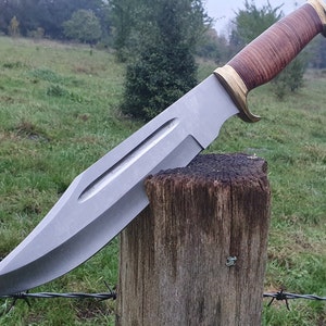 Puede incluir: Un cuchillo de caza grande, de color plateado, con mango de cuero marrón y guarda de latón. El cuchillo está apoyado en un poste de madera con alambre de púas en primer plano.