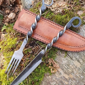 Puede incluir: Dos brochetas de metal forjadas a mano con un diseño retorcido y asas de bucle, junto con un tenedor a juego y una funda de cuero. El cuchillo tiene una hoja larga y curvada. Los artículos descansan sobre musgo verde y una superficie de madera envejecida.