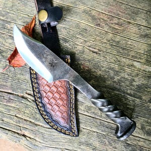 Puede incluir: Un cuchillo forjado a mano con un mango retorcido y una funda de cuero. La hoja es plateada y el mango es de metal oscuro. La funda es de cuero marrón con un patrón tejido y costuras amarillas. Una estrella está grabada en la hoja.