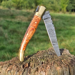 Peut inclure: Un couteau pliant avec un manche en bois et une lame en acier à motifs. Le manche est de couleur marron clair avec des accents en laiton. La lame est partiellement ouverte, révélant un motif tourbillonnant unique. Le couteau repose sur un morceau de bois.