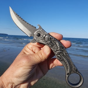 Op de afbeelding: Een zilveren zakmes met een drakenontwerp op het handvat. Het mes is gedeeltelijk open en het mes wordt in een hand gehouden.