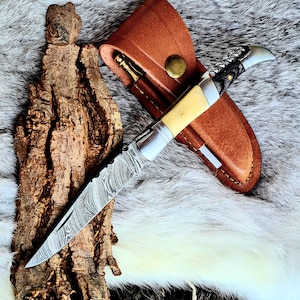 Könnte beinhalten: Ein Klappmesser mit einer Klinge aus Damaststahl und einem cremefarbenen Griff, zusammen mit einer braunen Lederscheide. Das Messer ist geöffnet und zeigt seine gemusterte Klinge. Ein Korkenziehermesser ist ebenfalls sichtbar. Die Scheide hat einen Messingknopf.