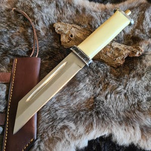 Può includere: Un coltello con manico bianco, lama argentata e fodero in pelle nera. Il coltello è appoggiato su una superficie di pelliccia bianca.