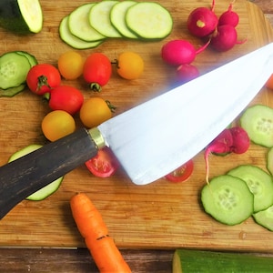 Può includere: Un grande coltello da chef affilato con manico in legno, appoggiato su un tagliere di legno. Il tagliere è circondato da diverse verdure, tra cui cetrioli, ravanelli, carote e pomodori.