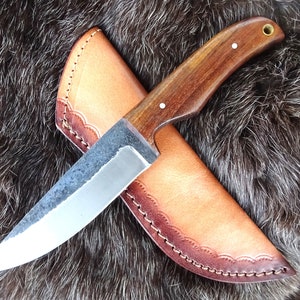 Puede incluir: Un cuchillo artesanal con mango de madera oscura y hoja plateada, presentado en una funda de cuero marrón claro. La funda tiene un borde festoneado decorativo y está cosida con hilo marrón. El cuchillo se muestra sobre un fondo oscuro y texturizado.