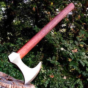 Op de afbeelding: Een handgesmeed bijl met een houten steel. De bijlkop is zilverkleurig met een afgeronde kling en een licht gebogen rand. De steel is van donker roodbruin hout.