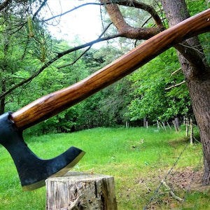 May include: A black axe with a wooden handle, resting on a tree stump in a grassy field. The axe has a curved blade and a sharp point. The handle is made of wood and has a natural finish.