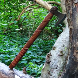 Può includere: Un'ascia con manico in legno marrone, avvolto in pelle, e lama curva in metallo grigio scuro. L'ascia è appoggiata su un tronco d'albero in un ambiente boschivo.