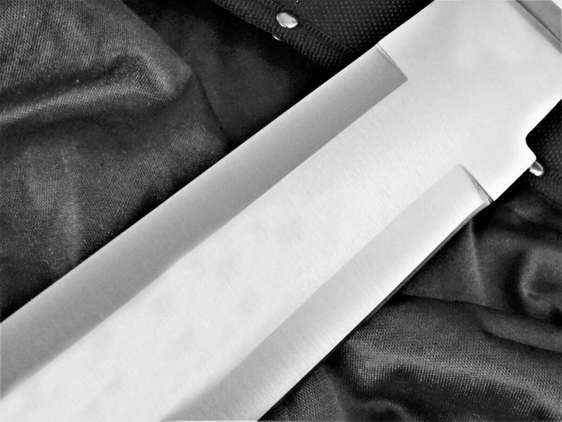 May include: A close-up of a silver knife blade with a satin finish. The blade is partially visible against a black fabric background.
