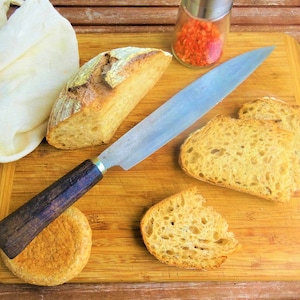 Puede incluir: Una tabla de cortar de madera con una hogaza de pan, un cuchillo y varias rebanadas de pan. El pan es de color marrón claro y tiene una corteza crujiente. El cuchillo es plateado con un mango de madera marrón. La tabla de cortar es de madera marrón claro.