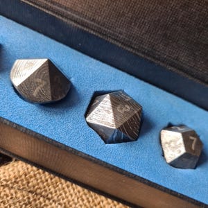Pode incluir: Conjunto de três dados geométricos com padrões gravados e numeração branca, exibidos numa caixa forrada a azul. Os dados têm uma aparência metálica, com texturas visíveis e arestas vivas. Os números 7 e 3 são visíveis.