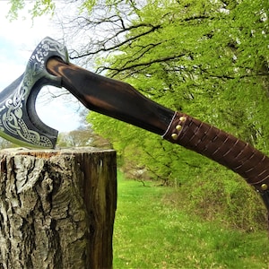 Könnte beinhalten: Eine handgeschmiedete Axt mit einem lederbezogenen Stiel und aufwendigen Metallarbeiten. Der Axtkopf zeigt ein keltisches Knotendesign und ist auf einem Holzstiel montiert.