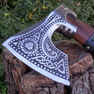 Könnte beinhalten: Eine silberne Axt mit einem Holzstiel und aufwendigen, geätzten Designs, darunter ein keltischer Knoten und ein Valknut-Symbol.