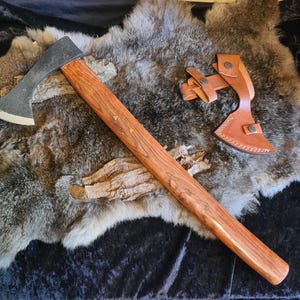 Puede incluir: Un hacha negra y marrón con un mango de madera y una funda de cuero. El hacha está sobre una piel gris y blanca.