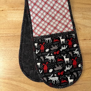 Könnte beinhalten: Gesteppter Ofenhandschuh mit einem schwarzen, roten und weißen Muster. Der obere Teil hat ein rot-weißes Karomuster, während der untere Teil einen schwarzen Hintergrund mit Illustrationen von Hirschen, Elchen und Kaninchen hat. Der Handschuh ist mit schwarzem Stoff eingefasst.